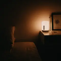 Łóżko z poduszką w półmroku. Lampka nocna rozprasza mrok, łagodząc objawy nyktofobii. Na stoliku butelka i obrazek.
