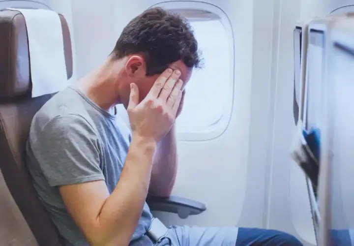Mężczyzna w samolocie z rękami na twarzy, wyraźnie cierpiący na jet lag po długiej podróży.