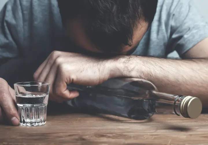Zmęczony mężczyzna opiera głowę na butelce alkoholu, obok stoi kieliszek wody. To symboliczny obraz odstawienia alkoholu i walki o trzeźwość.