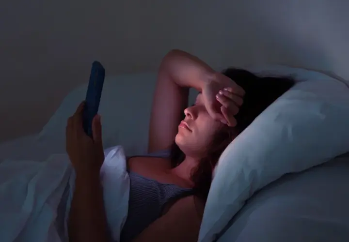 Kobieta leży w łóżku, przeglądając telefon w środku nocy. Jej ręka zasłania oczy, oznaka bezsenności.