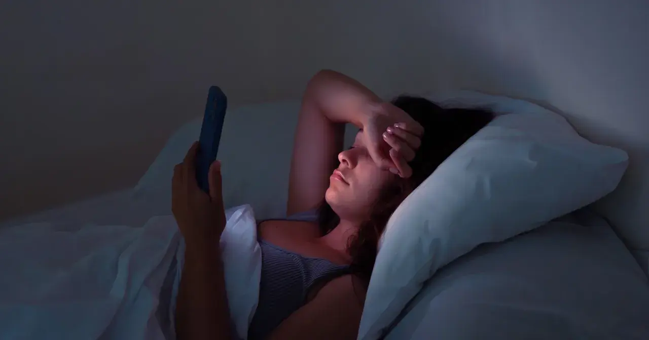 Kobieta leży w łóżku, przeglądając telefon w środku nocy. Jej ręka zasłania oczy, oznaka bezsenności.