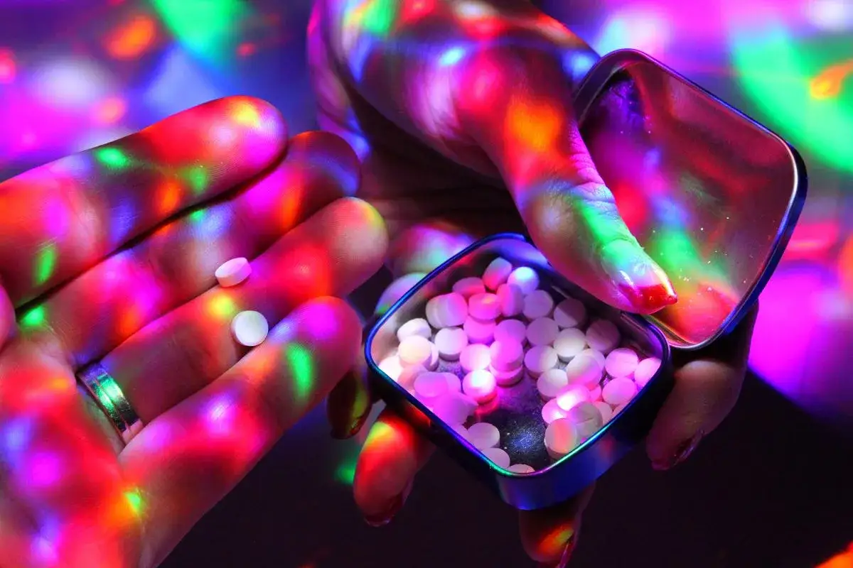 Dłonie oświetlone kolorowymi światłami, trzymające małe białe tabletki, przypominające te zawierające mdma.